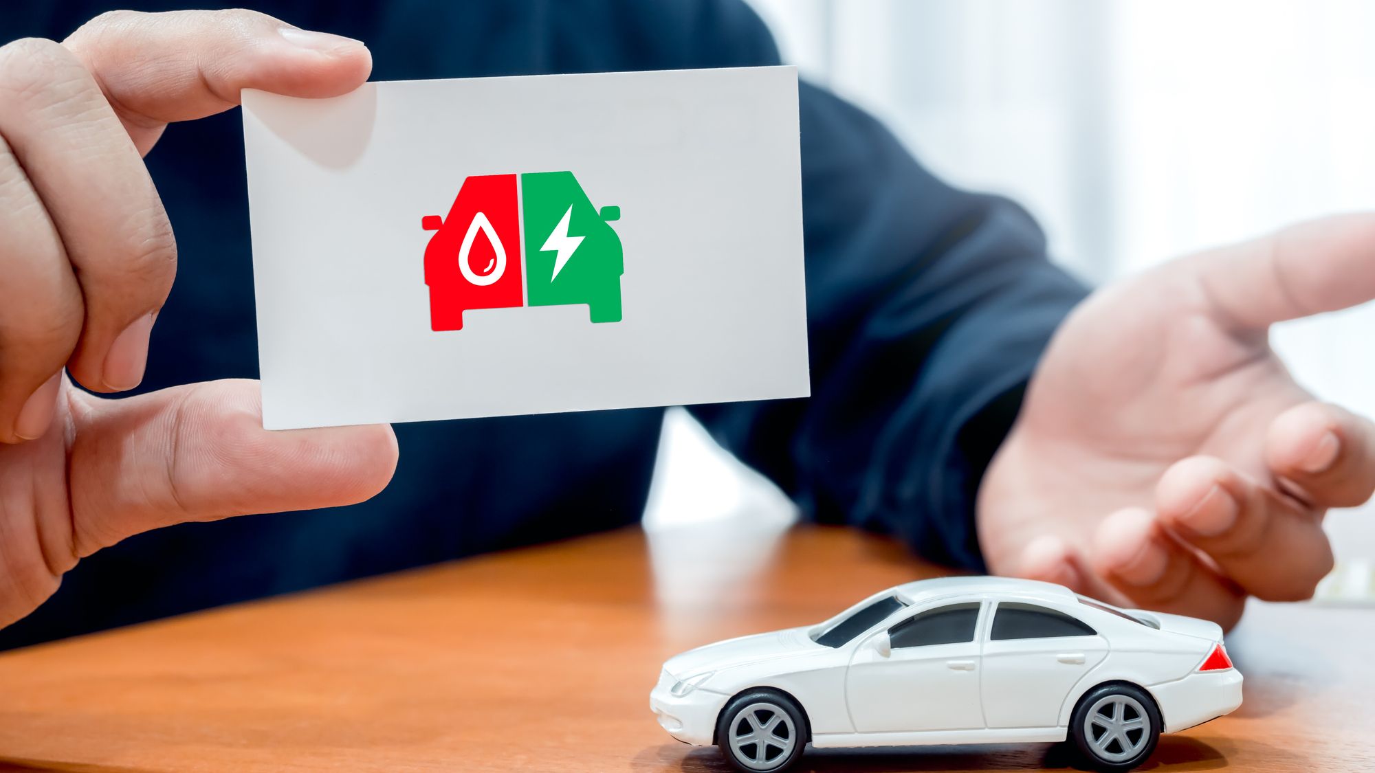 A hand holding a card with electric and fuel engine icons next to a toy car, representing a consumer's choice between powertrains.