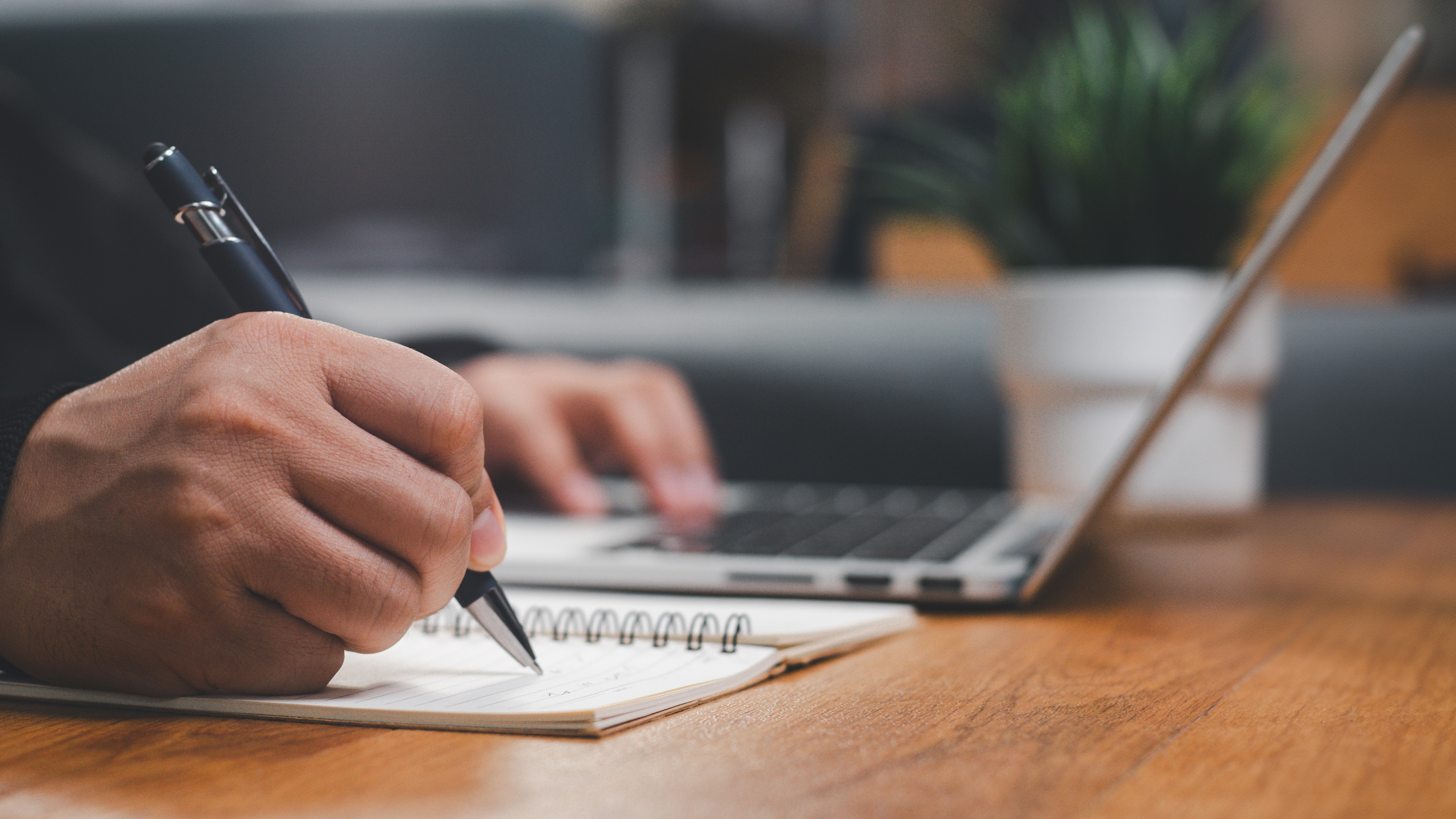 A man taking notes in a spiral notebook while using a laptop, focusing on detailed financial work.