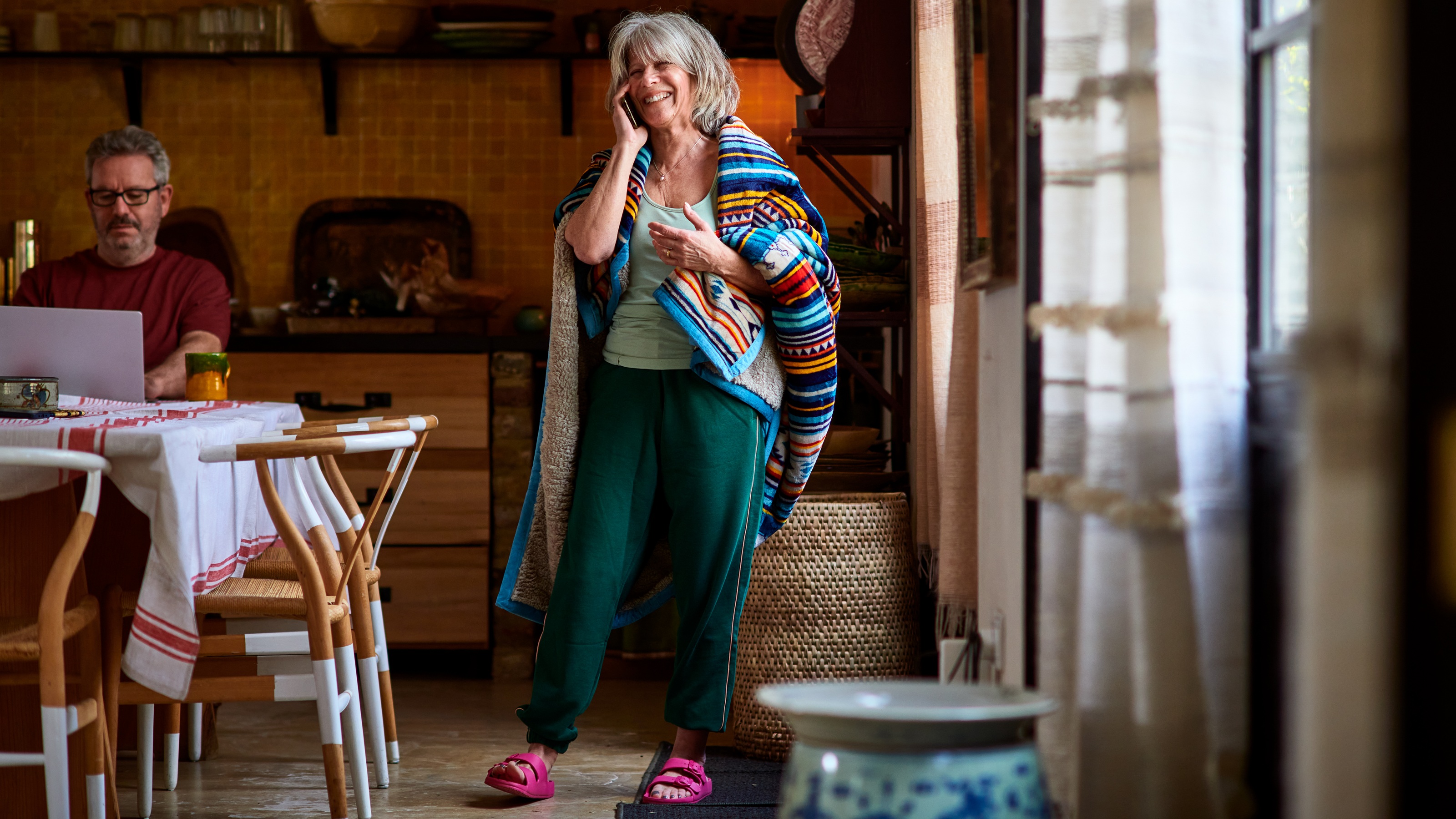 An older couple in a comfortable kitchen setting, the woman smiling on the phone while the man works on a laptop, representing financial peace of mind.
