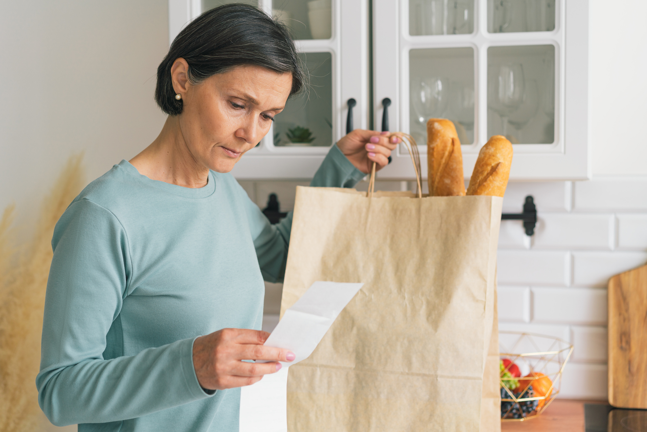 A woman thoughtfully reviewing a long grocery receipt in a bright kitchen setting.