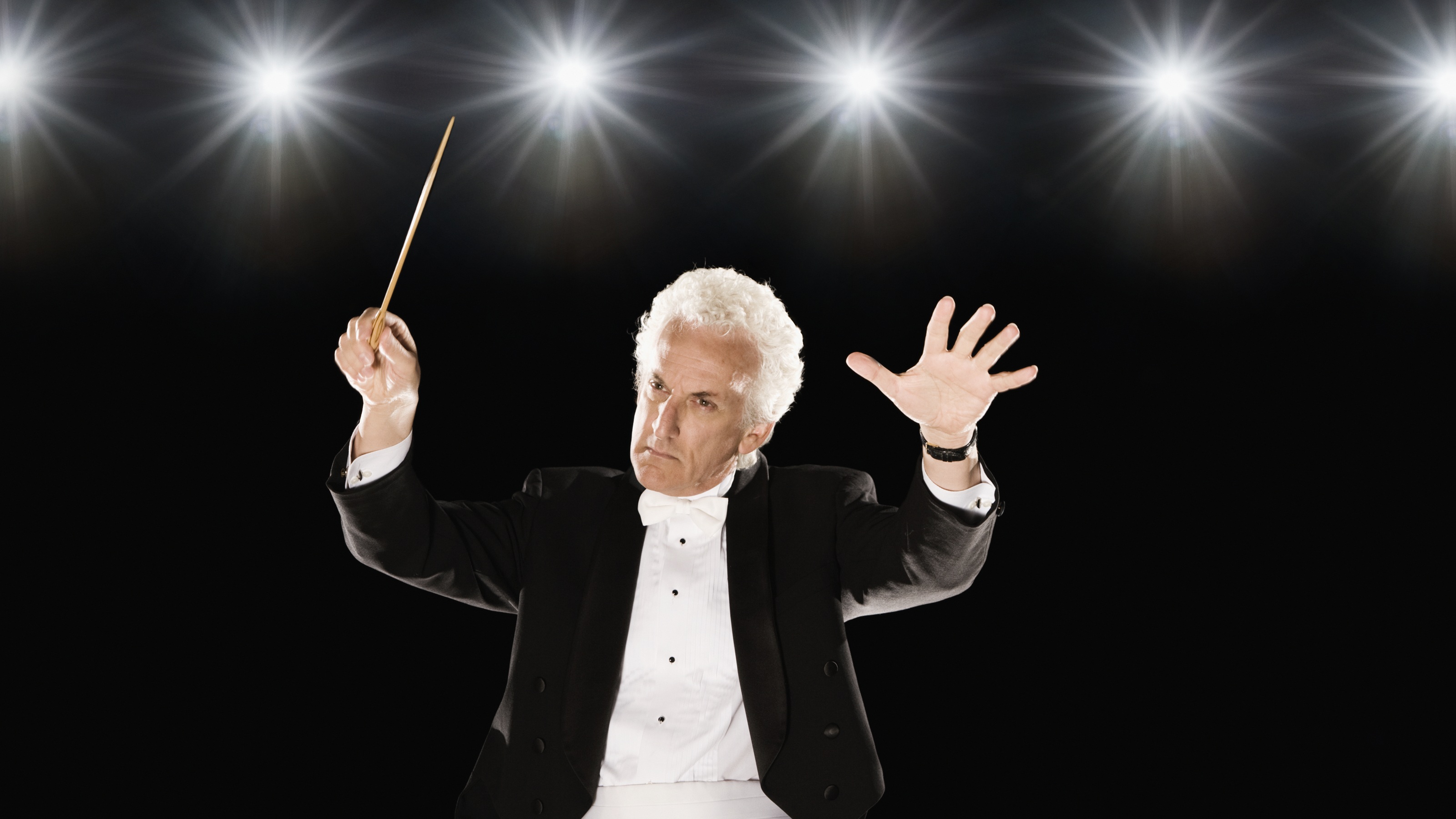 A male conductor in a tuxedo gesturing as if leading an orchestral performance.